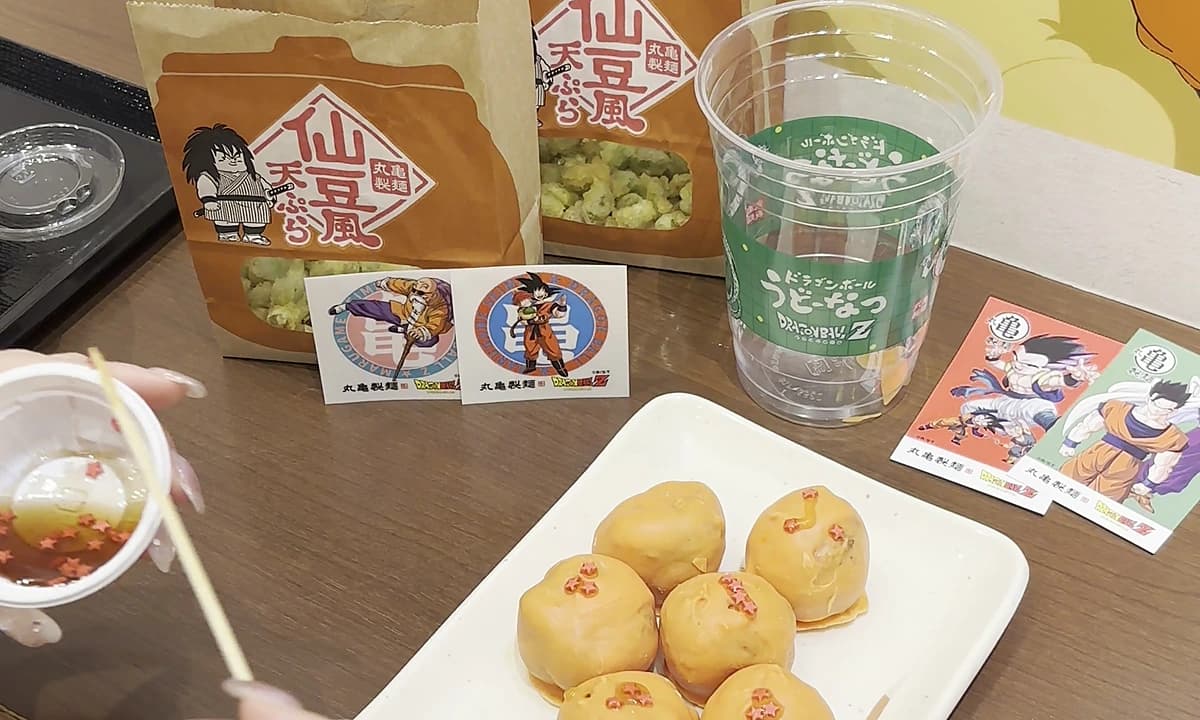 Dragon Ball Z collab dining table at Marugame Seimen with Senzu-style tempura takeout bags, character udon tickets, themed cup, takoyaki plate, painted Goku mural backdrop