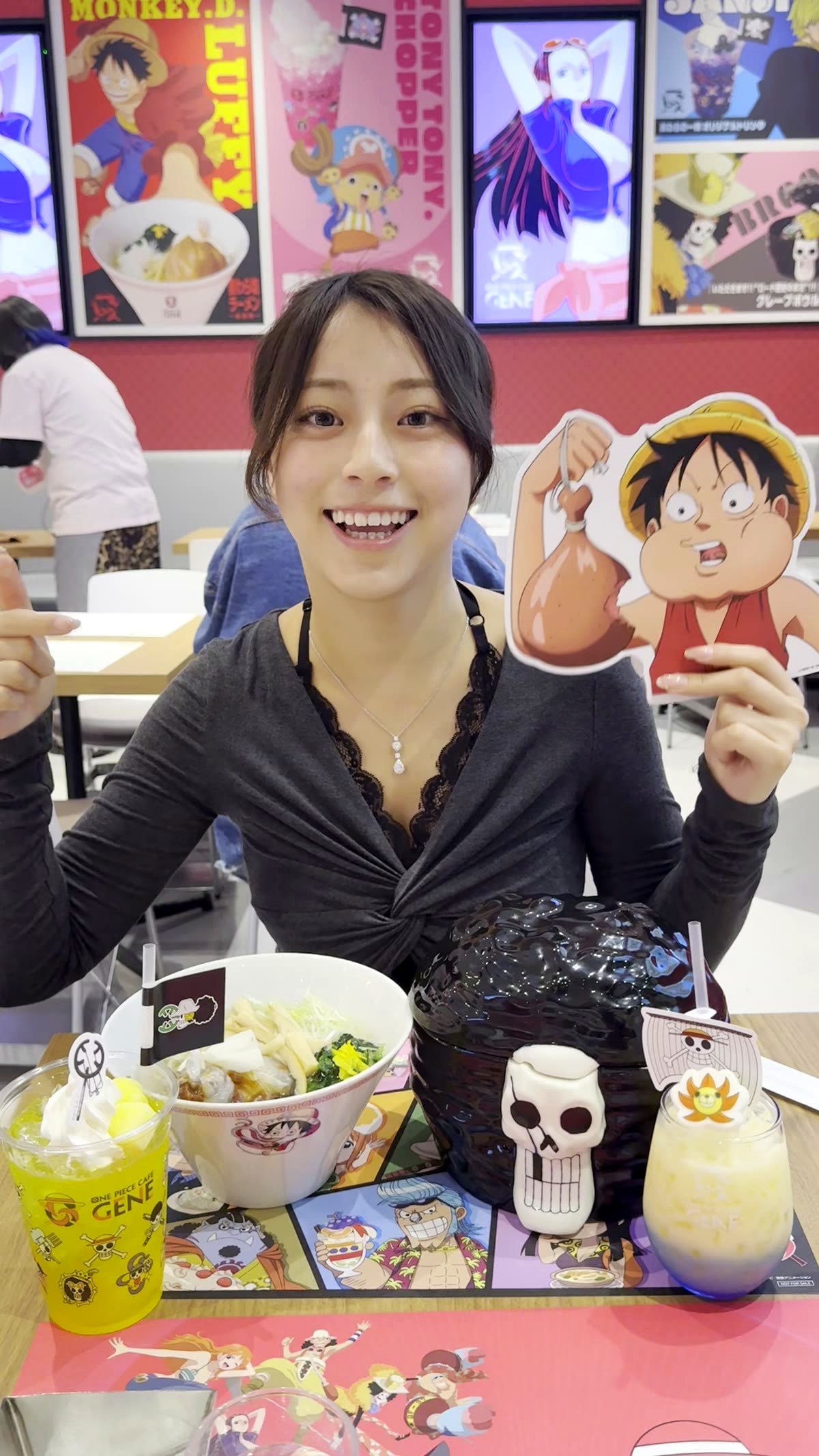 Moe from Japan Pop Now with full table spread at One Piece Cafe GENE Shibuya — skull bowl ramen, crepe bowl, two character drinks, Luffy standee, and Straw Hat Crew placemat